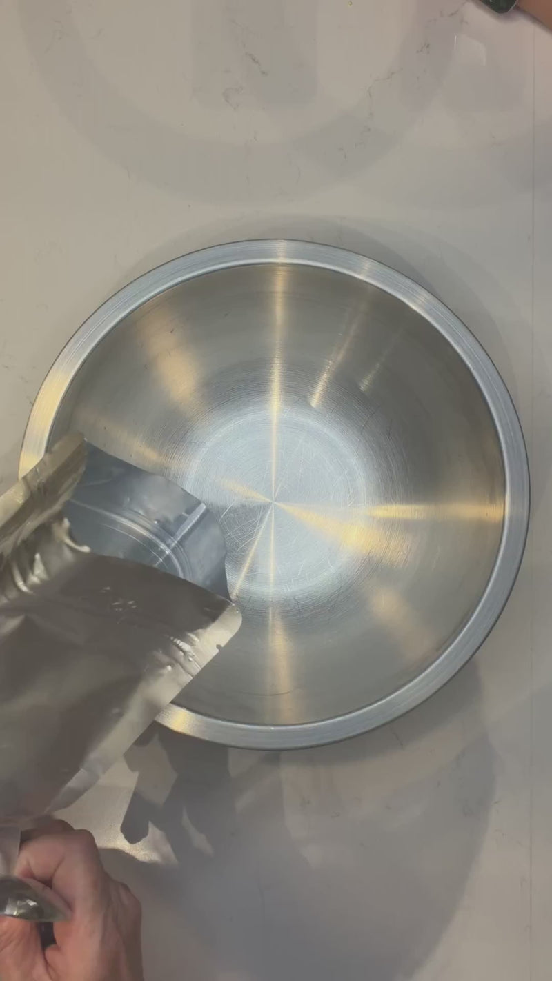 A hand pouring the dry pouch of The Everything cookie kit into a steel mixing bowl